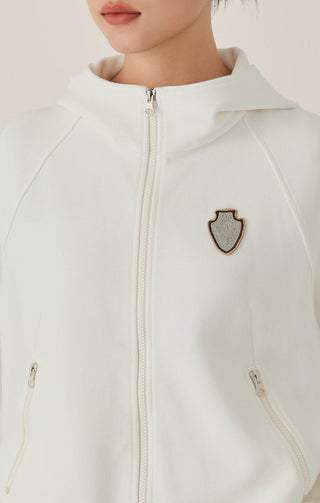 Close-up of white zip hoodie: center front zipper and beaded shield crest badge.