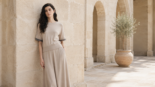 A model wearing a neutral knit top and flowing wide-leg skirt in a sunlit Mediterranean stone courtyard